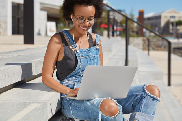 妇女穿着时髦的破烂工作服 打视频电话 使用现代笔记本电脑和耳机 坐在楼梯旁 听有声书 积极微笑的满意黑人女青年户外镜头笔记本电脑破布遥控