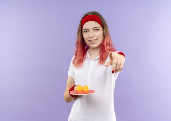 球戴着头巾 拿着乒乓球拍和球的年轻运动女性 手指着相机微笑着站在紫色的墙上抱着手指球拍
