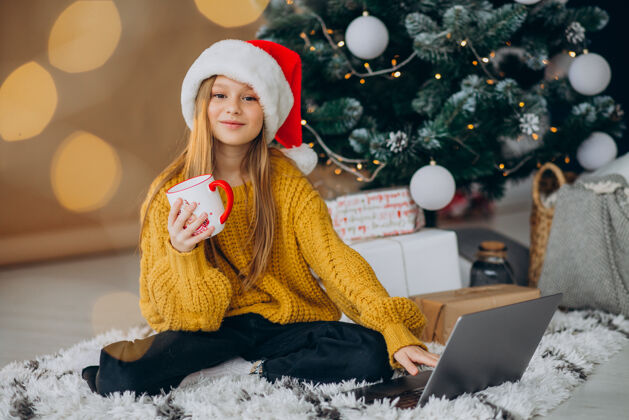 在线可爱的女孩在圣诞树旁用电脑网上商店圣诞销售科技