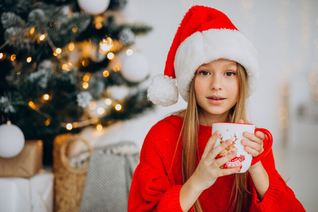 人在圣诞树旁喝可可的女孩看礼物红帽子