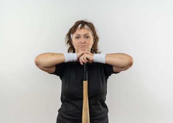 运动装身穿黑色t恤 头戴棒球棒的中年运动女性站在白色的墙壁上不高兴棒球头带女