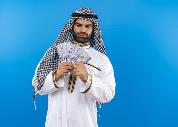 显示身着传统服装的阿拉伯男子站在蓝色的墙上 表情自信严肃 看上去很有钱表达信心站