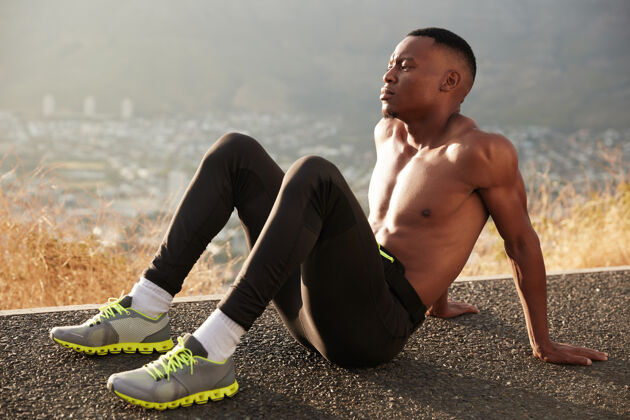 自信佩西的横观黑色男性斜靠在手上 坐在马路上 有着肌肉发达的身体 穿着运动裤 运动鞋 白袜子在户外慢跑运动鞋沥青户外