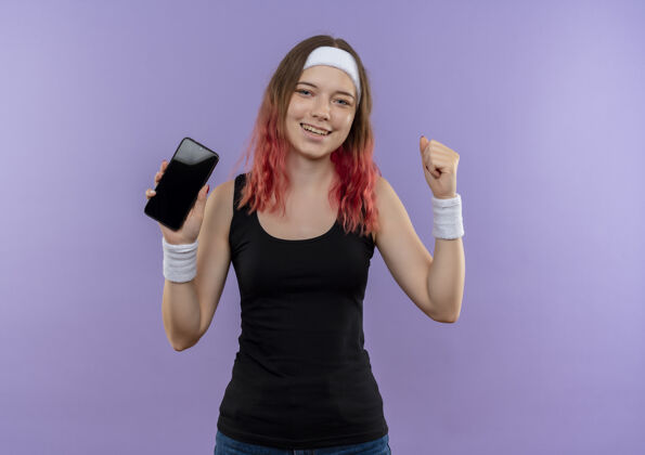 人身着运动服的年轻健身女士展示智能手机握紧拳头 开心地微笑着 站在紫色的墙上离开了表演运动移动