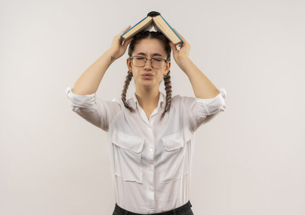 抱戴着眼镜 梳着辫子 穿着白衬衫的年轻女学生头上举着一本打开的书 站在白墙上显得很失望女孩书衬衫