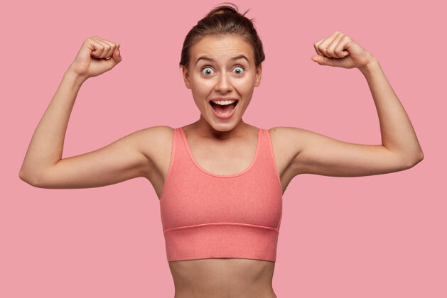 皱眉图为强壮开朗的年轻女运动员在健身房锻炼肌肉 穿着与墙壁同色调的休闲上衣 拥有健康完美的身体 令人惊叹运动理念衣服时尚二头肌