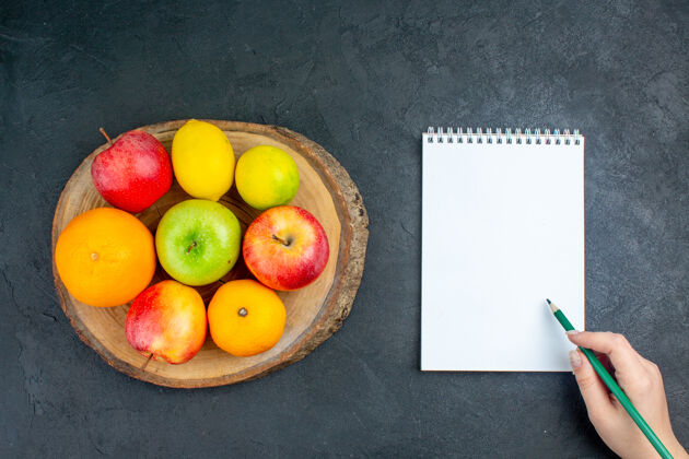 苹果俯视图苹果柠檬橘子木板笔记本铅笔在女性手中黑暗的表面苹果柠檬纸板