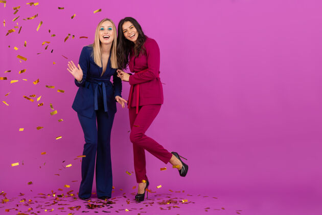 兴奋两位魅力四射的女士在紫罗兰色的墙上欢庆新年 穿着紫色和蓝色的时尚多彩的晚礼服 朋友们在一起玩得很开心 时尚潮流 金色的五彩纸屑派对气氛夏娃庆典优雅