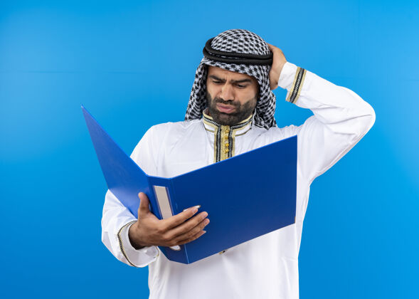 阿拉伯语阿拉伯商人穿着传统的衣服拿着打开的文件夹 看着它困惑地用手顶着头站在蓝色的墙上手穿寻找