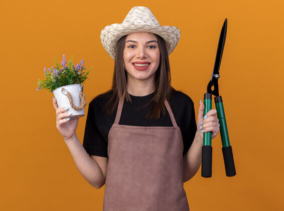 花盆微笑着美丽的白人女园丁戴着园艺帽拿着花盆和园艺剪刀隔离在橙色的墙上 有复制空间园艺花园漂亮