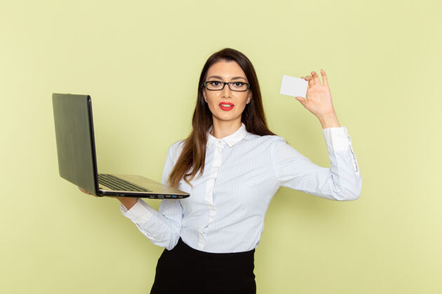 微笑穿着白衬衫和黑裙子的女上班族在浅绿色墙上使用笔记本电脑和名片的前视图女前面拿着