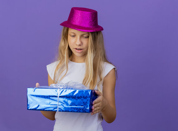 生日漂亮的小女孩戴着节日礼帽拿着礼盒看着它惊讶不已 生日派对的概念站在紫色的背景上紫色聚会小