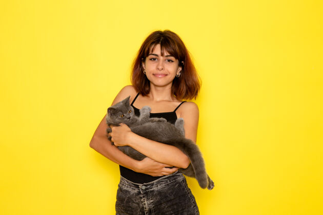 情感身穿黑色衬衫和灰色牛仔裤的年轻女性的正面视图 黄色墙上抱着灰色的猫女孩前面姿势