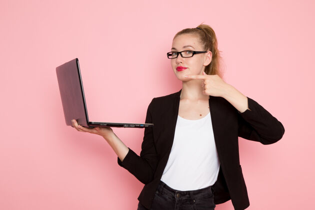 办公室穿着黑色夹克的女上班族在浅粉色墙上拿着笔记本电脑的正视图人笔记本电脑忙碌