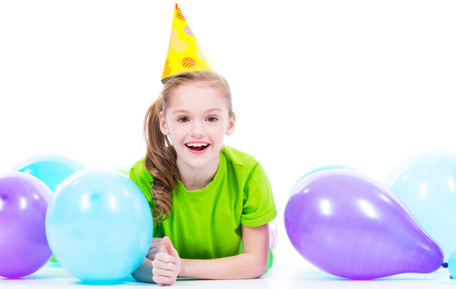 欢呼穿着绿色t恤的快乐微笑女孩躺在地板上 带着五颜六色的气球-隔离在白色的地板上竖起大拇指白种人庆祝