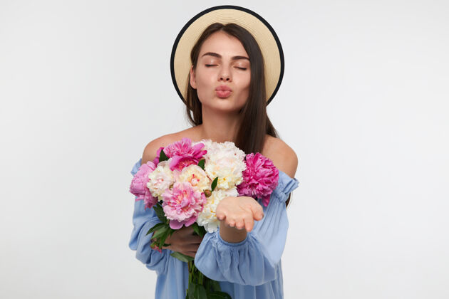 面部一个漂亮漂亮的女孩的肖像 长着深褐色的长发 戴着帽子 穿着蓝色的裙子 手持一束鲜花 送上一个吻 孤立地站在白色的墙上花长发模特