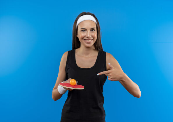 乒乓球微笑的年轻漂亮的运动女孩戴着头带和腕带乒乓球拍与球 并指着它在蓝色的空间蓝色运动年轻