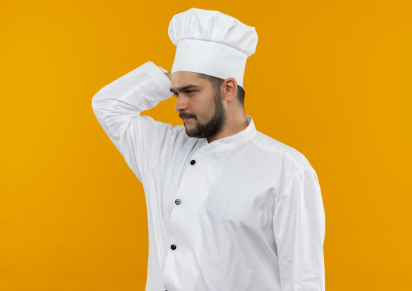 厨师体贴的年轻男厨师身着厨师制服 把手放在脑后 看着橙色空间上孤立的一面橘子手烹饪