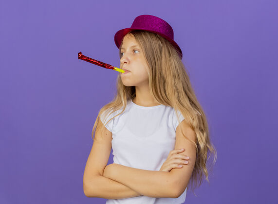 帽子得罪了漂亮的小女孩在节日帽子吹哨子 生日聚会的概念站在紫色的背景吹着漂亮小
