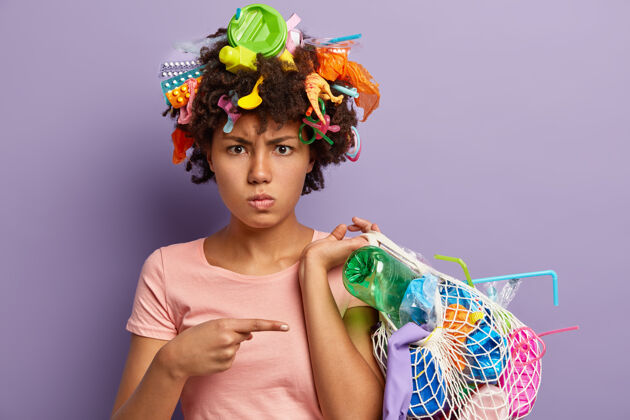垃圾不高兴的非洲裔美国妇女对滥用塑料感到愤怒的照片 指着收集垃圾的袋子 头上有垃圾 隔离在紫色的墙上不可回收的污染概念关怀压力浪费