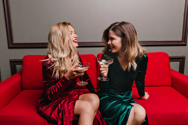 优雅好脾气的女孩喝酒聊天室内照片 高兴的女士坐在红色沙发上 戴着酒杯卷发微笑朋友