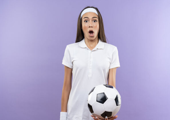 足球惊讶漂亮的运动女孩戴着头带和手环拿着足球隔离在紫色的空间穿着紫色运动