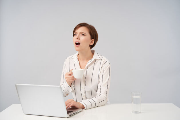 水年轻的黑发短发女人 随意的发型 一边看一边喊着什么 一边喝茶一边把手放在笔记本电脑的键盘上 孤零零地站在白色的地板上深色坐着办公室