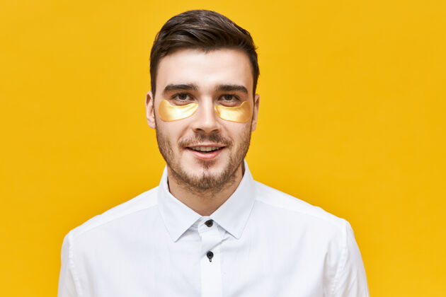 卫生时尚的未剃胡子的年轻白人男性戴着眼罩 以解决脱水和黑眼圈 因为紧张的生活方式 摆出黄色墙壁 贴片头头发