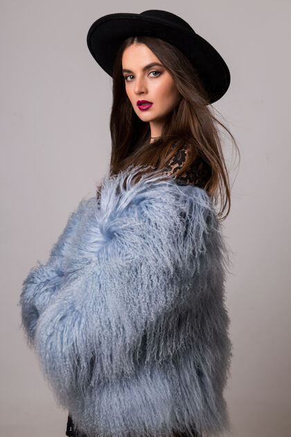 最好穿着时髦的冬季蓬松的蓝色外套和黑色帽子的漂亮女人的时尚肖像在明亮的灰色墙上摆姿势年轻冷女人