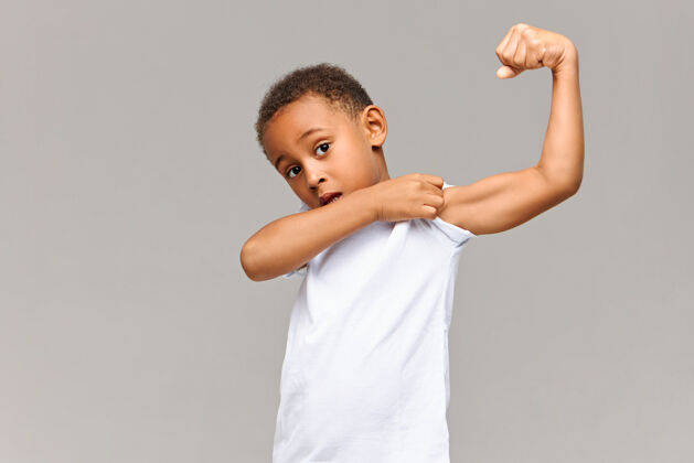 孩子看看我的二头肌有趣的非裔美国人在休闲白色t恤的照片摆在灰色墙拉袖子孤立 显示他紧张的手臂肌肉童年 健身和运动的概念肌肉孩子休闲