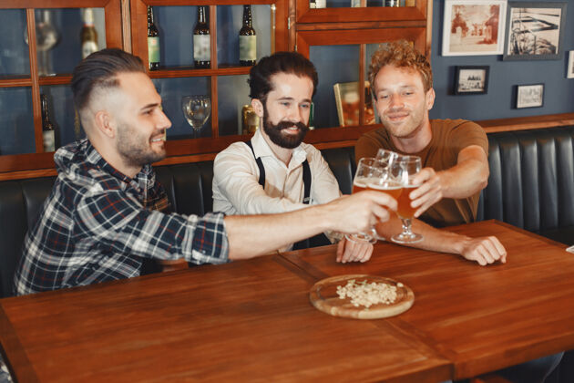饮酒和最好的朋友见面三个穿着休闲服的快乐的年轻人一起坐在酒吧里聊天喝啤酒坐着微笑休闲