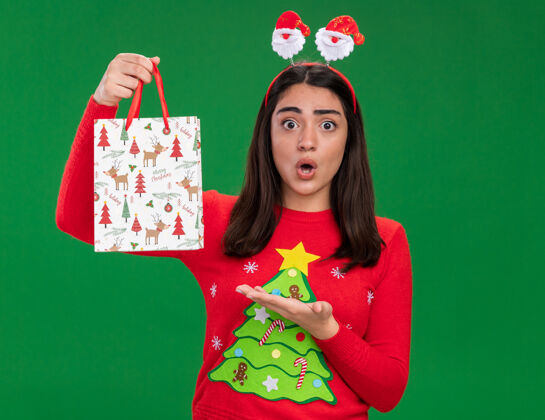 点震惊的年轻白人女孩与圣诞老人头带举行和点在纸礼品袋隔离在绿色背景与复制空间快乐圣诞快乐礼物