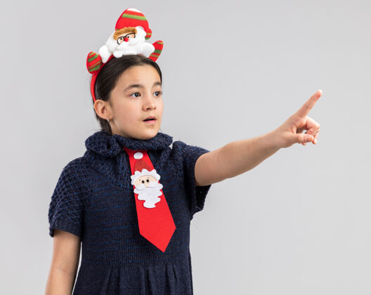 圣诞小女孩穿着针织连衣裙 头上系着一条红色领带 带着滑稽的圣诞边 用食指指着旁边的东西穿边缘索引