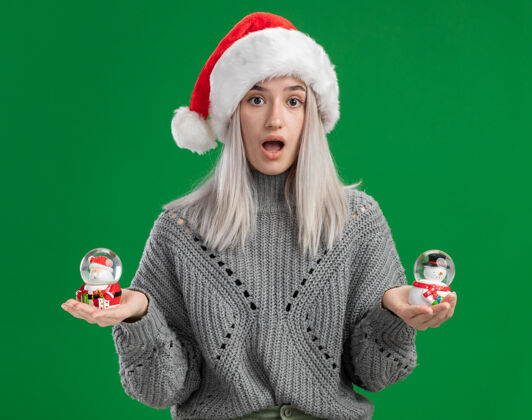 抱着年轻的金发女郎穿着冬天的毛衣 戴着圣诞帽 手里拿着圣诞玩具雪球 站在绿色的背景上惊讶地看着相机毛衣圣诞老人地球仪