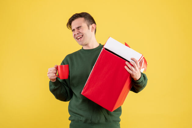 快乐的人正面图：穿着绿色毛衣的快乐男人拿着大礼物 红色的杯子站在黄色的椅子上大礼物毛衣