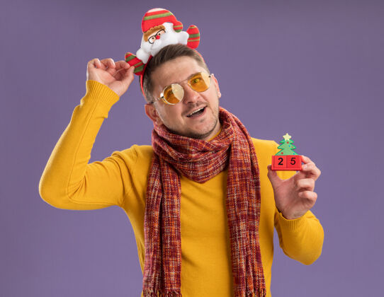 数字快乐快乐的年轻人穿着黄色高领毛衣 戴着温暖的围巾和眼镜 戴着滑稽的帽沿 头上戴着圣诞老人 在紫色的墙上展示着25号玩具立方体穿着圣诞老人头