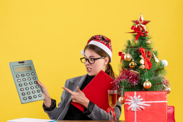 桌子前视图漫步的女孩戴着圣诞帽坐在桌子旁看着计算器圣诞树和礼物鸡尾酒流浪鸡尾酒笔记本电脑