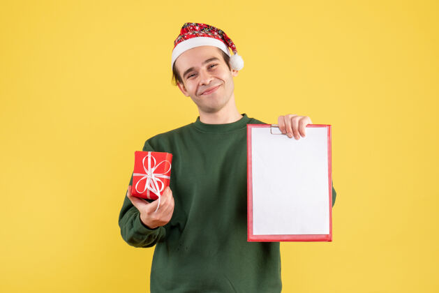 成人正面图：穿着绿色毛衣的年轻人拿着剪贴板和礼物站在黄色的地板上男人站着帅哥