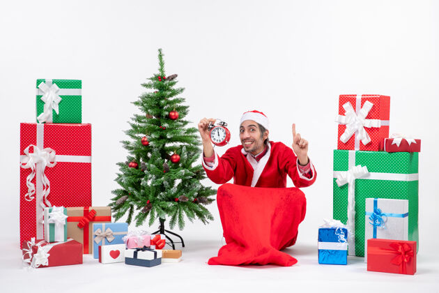 坐着圣诞老人展示了一个坐在地上 在礼物和圣诞树旁展示时钟的圣诞老人装饰礼物树