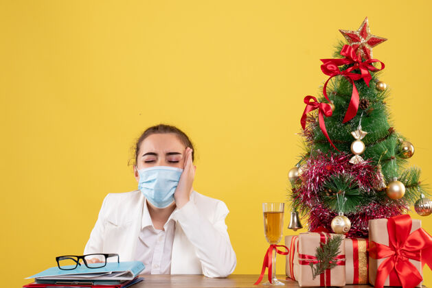 面部正面图女医生戴着防护面具坐在黄色背景上 戴着圣诞树和礼盒坐着女医生实验室
