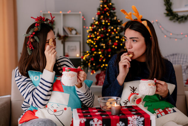 头带带着冬青花环和驯鹿头带的快乐的年轻女孩们拿着杯子 坐在扶手椅上吃饼干 在家里享受圣诞节时光圣诞快乐快乐年轻