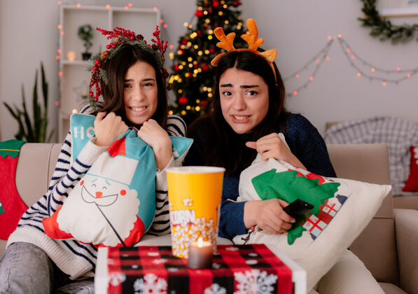 扶手椅带着冬青花环和驯鹿头带的年轻女孩们在家里坐在扶手椅上抱着枕头看电视驯鹿坐着圣诞快乐