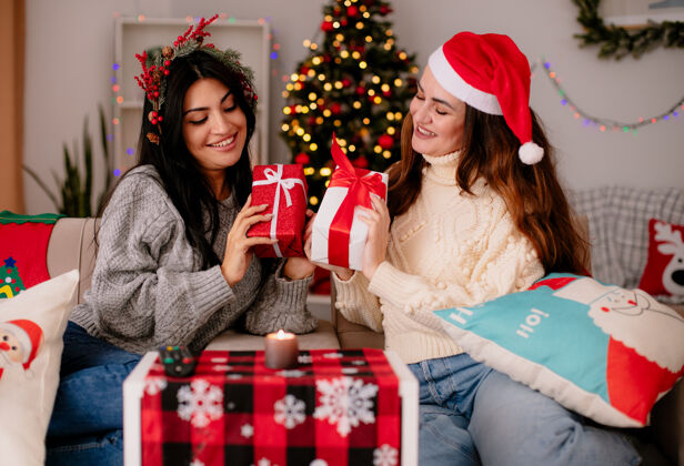 圣诞快乐带着圣诞帽和冬青花环的年轻女孩们微笑着拿着礼物盒坐在扶手椅上 在家里享受圣诞节时光年轻享受扶手椅