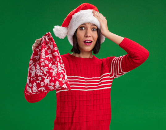 保持令人印象深刻的年轻女孩戴着圣诞帽拿着圣诞礼物袋手放在头上隔离在绿色的墙上圣诞节年轻麻袋