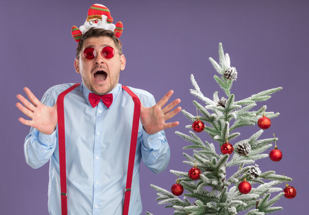 蝴蝶结戴吊带领结的年轻人站在圣诞树旁 戴着圣诞老人和红眼镜 张着大大的嘴站在紫色的墙上 惊讶地看着站立惊奇开放