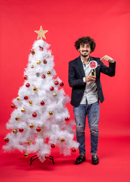 肖像兴奋的年轻人拿着钟和一杯酒 站在红色的圣诞树旁葡萄酒时钟年轻人