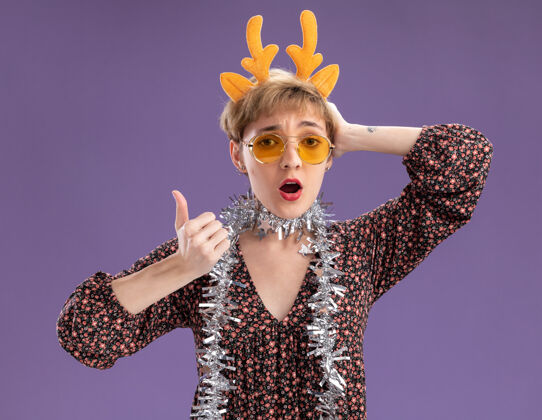 头带令人印象深刻的年轻漂亮女孩戴着驯鹿鹿角头带 脖子上戴着金箔花环 戴着眼镜 手放在头上 在紫色的墙上孤立地竖起大拇指年轻圣诞节保持