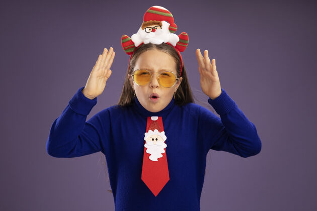 领带小女孩穿着蓝色高领毛衣 打着红色领带 头上戴着有趣的圣诞戒指 惊讶地举起双臂提高小搞笑