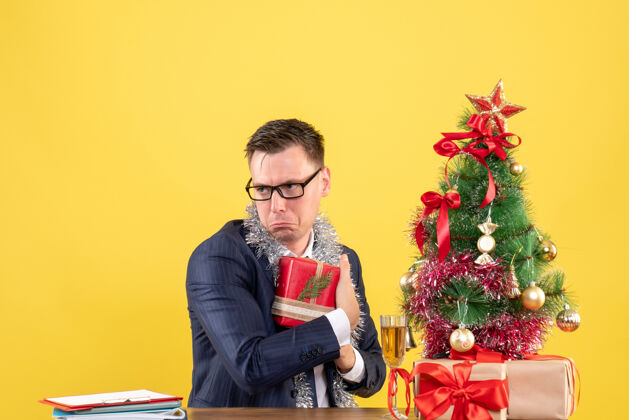 男人郁闷的男人把礼物藏在圣诞树旁的桌子上 礼物放在黄色的桌子上新郎树办公室