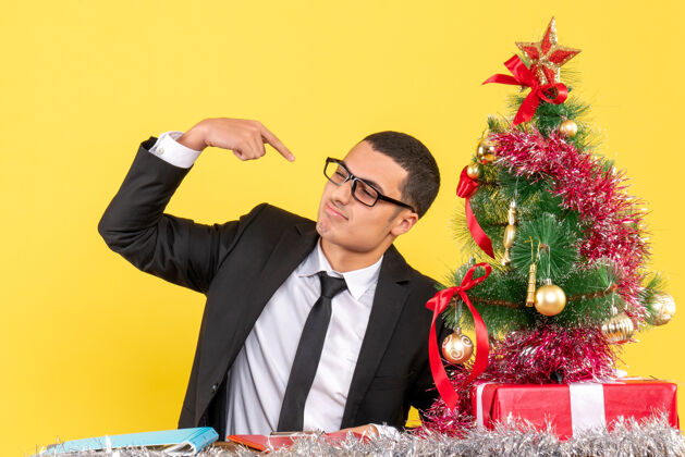 桌子正面图：一个穿着西装的年轻人坐在桌子旁 用手指着自己的圣诞树和礼物他自己礼物专业
